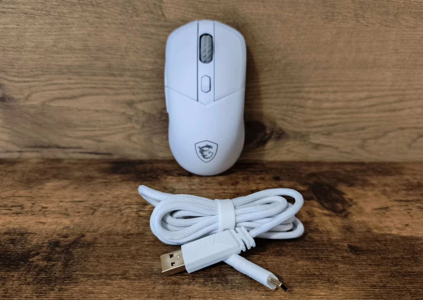 Biała mysz komputerowa z logo, ustawiona pionowo na drewnianym tle, poniżej biały przewód USB zakończony wtyczką USB-A i USB-C.