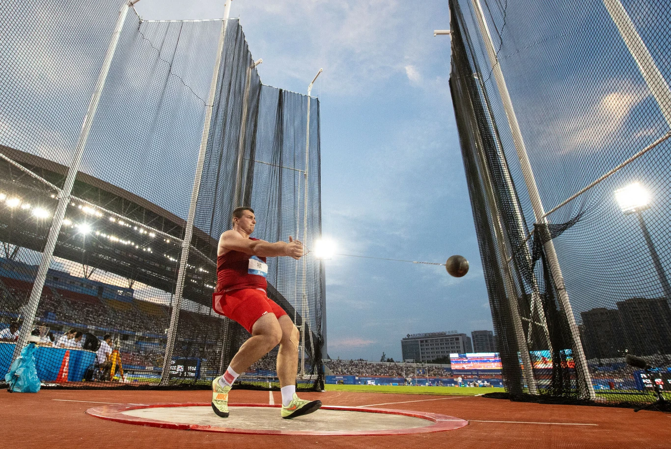 Lekkoatleta w czerwonym stroju wykonuje rzut młotem na stadionie lekkoatletycznym podczas zawodów, młot znajduje się w powietrzu, wokół widoczne są zabezpieczające siatki oraz trybuny z publicznością.