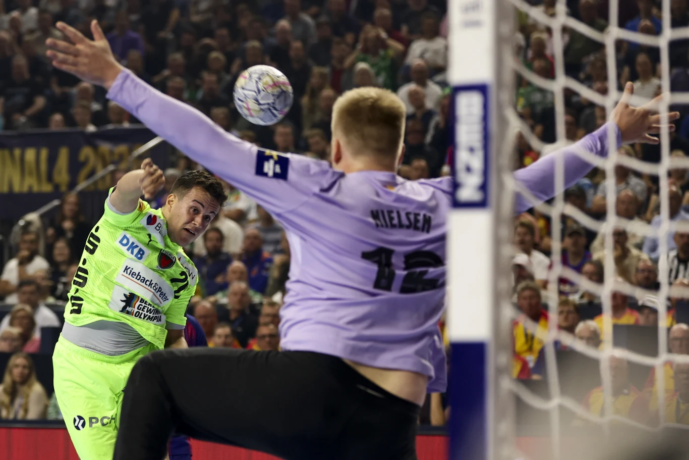 Piłkarz ręczny w jasnozielonym stroju wykonuje rzut na bramkę bronioną przez bramkarza w fioletowej koszulce z numerem 12 podczas meczu w hali sportowej wypełnionej kibicami.