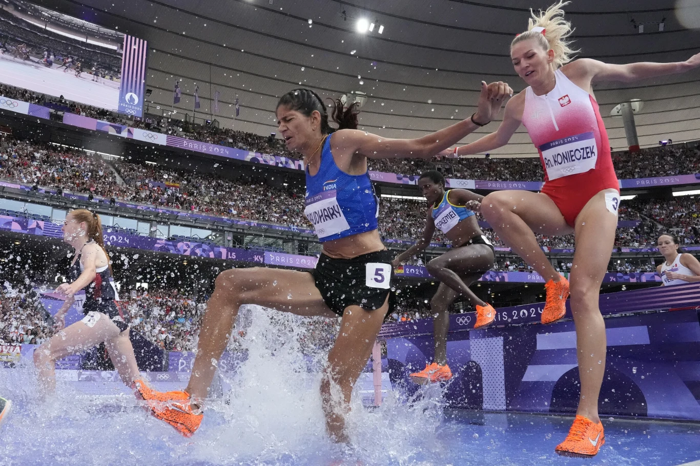 Grupa lekkoatletek w trakcie biegu z przeszkodami przeskakuje przez wodny rów, rozpryskując wodę podczas rywalizacji na stadionie olimpijskim.