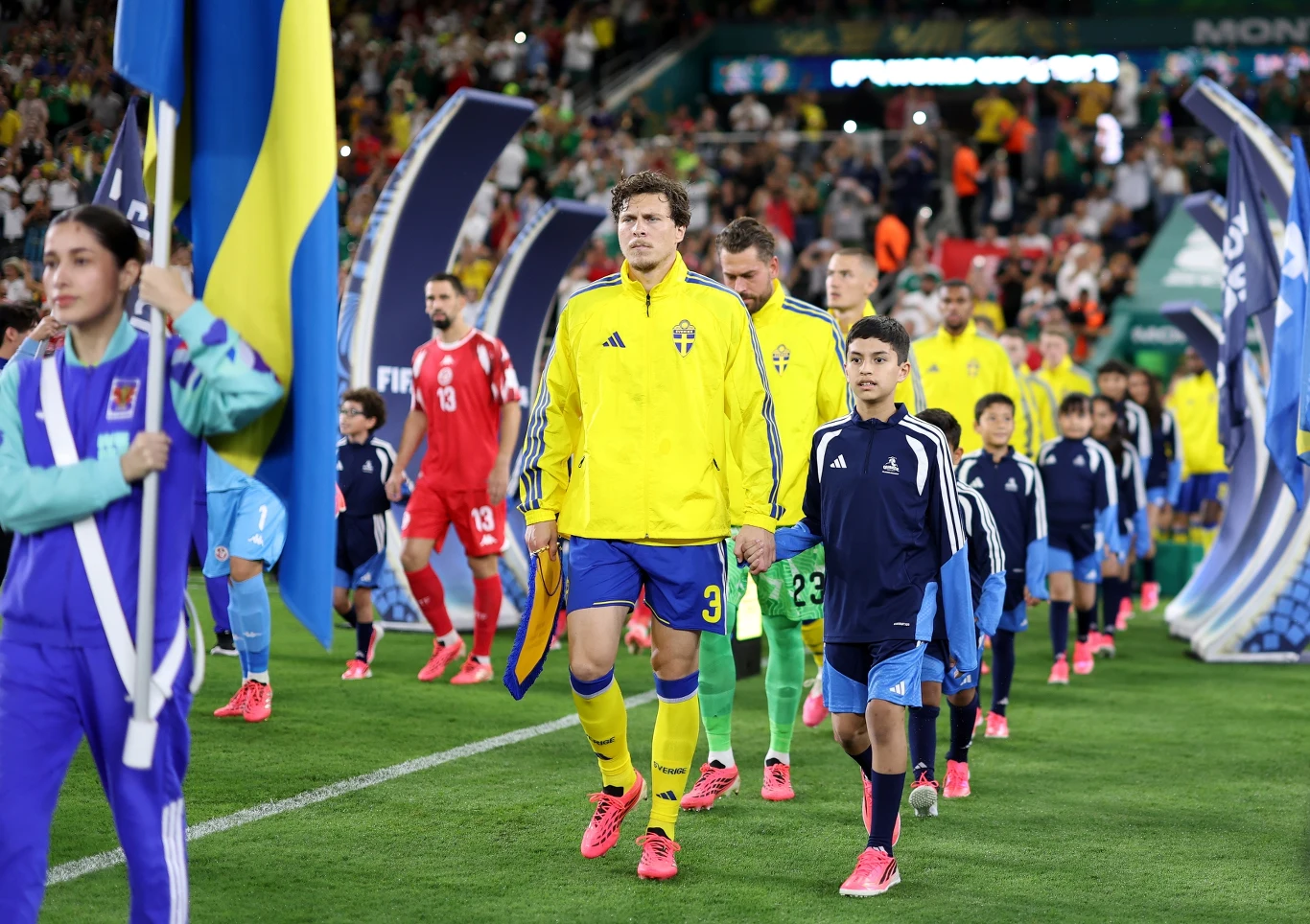 Piłkarze reprezentacji Szwecji w żółtych strojach oraz reprezentanci drużyny przeciwnej ubrani na czerwono wchodzą na murawę stadionu prowadzeni przez dziecięcych eskortujących i osoby z flagami, wokół widoczne są liczne flagi i tłum kibiców na trybunach.