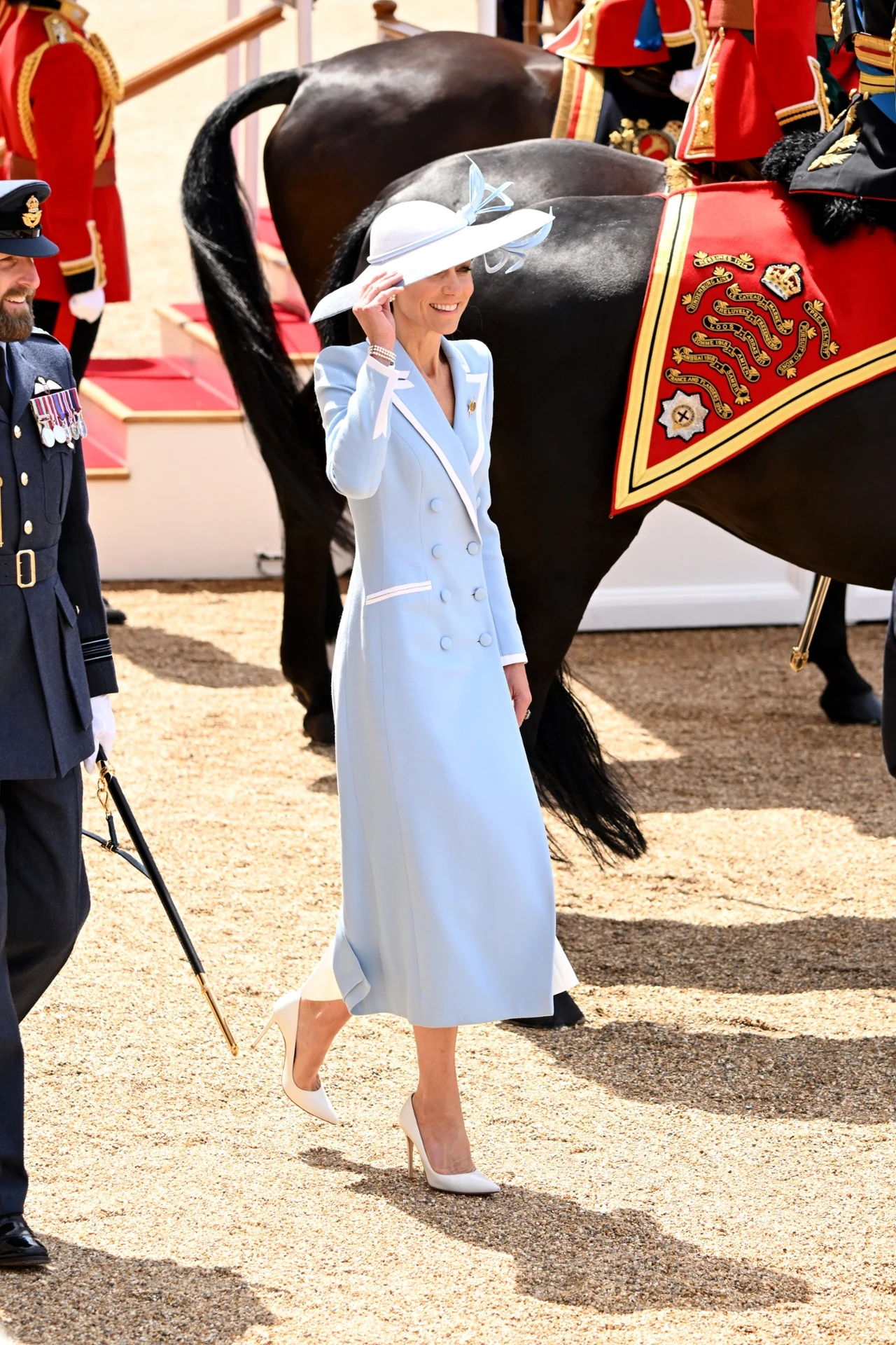 Kobieta w eleganckim, jasnoniebieskim płaszczu i dużym kapeluszu idzie obok czarnego konia z ozdobnym rzędem oraz mężczyzny w mundurze wojskowym.