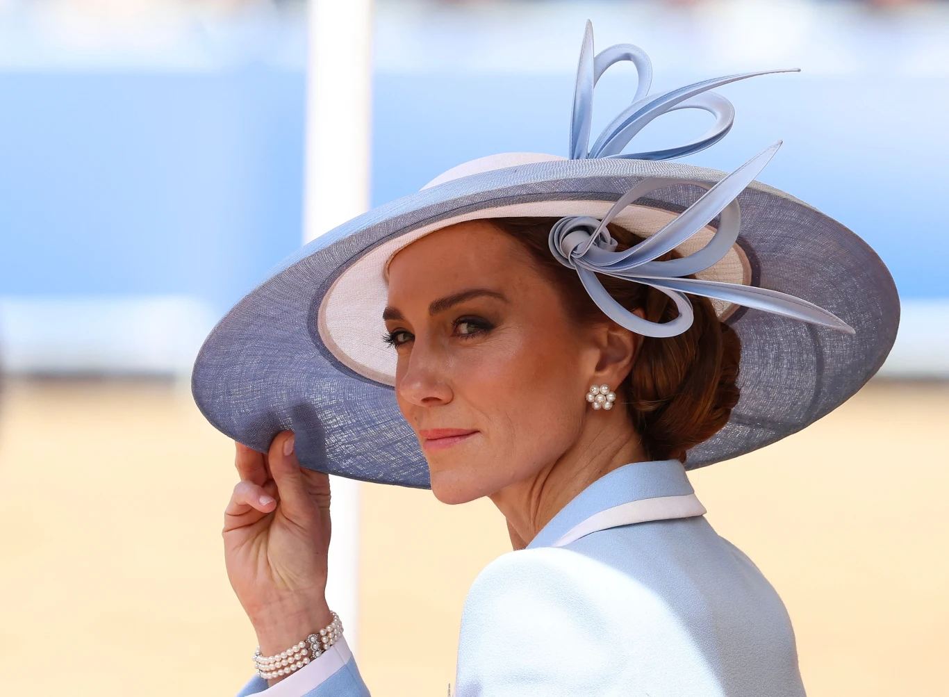 Kobieta w eleganckim, jasnoniebieskim kapeluszu z dekoracyjnym elementem, ubrana w jasny żakiet, nosząca perłowe kolczyki i bransoletkę, spogląda w stronę obiektywu.