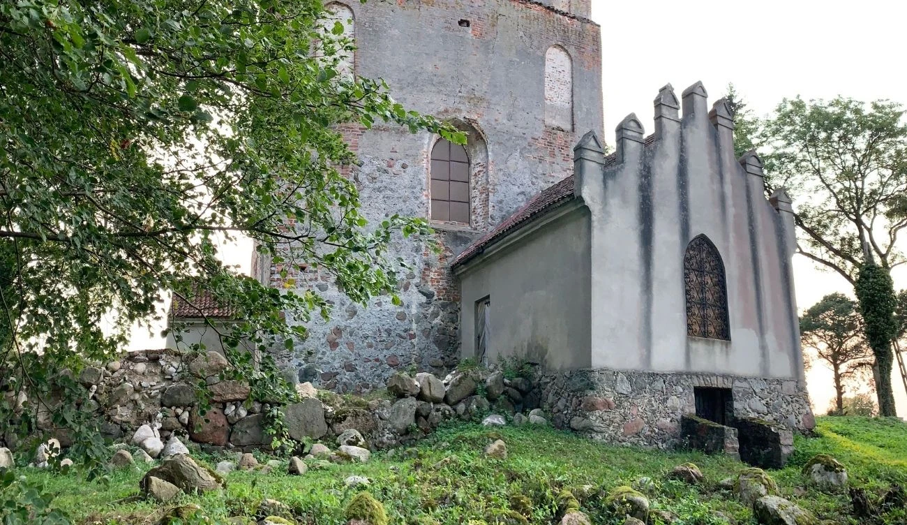 Kamienny budynek o surowych murach z gotyckimi elementami architektonicznymi, otoczony zielenią i fragmentami starych murów oraz głazów.