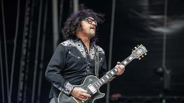 Mężczyzna z burzą ciemnych włosów i okularami grający na czarnej gitarze elektrycznej w trakcie koncertu na scenie, ubrany w czarną koszulę z białymi haftowanymi zdobieniami.