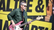 Mężczyzna w ciemnych okularach grający na czerwonym gitarze basowej podczas koncertu na tle dużego, żółtego banera z czarnymi napisami.
