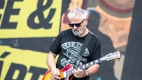Mężczyzna z siwymi włosami i brodą gra na gitarze elektrycznej, ubrany w czarną koszulkę z napisem i ciemne okulary przeciwsłoneczne, na tle dużych, żółto-czarnych liter.