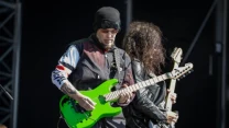 Dwóch gitarzystów grających na scenie plenerowej, z przodu mężczyzna w czarnej czapce i okularach na czole trzymający jasnozieloną gitarę elektryczną, obok niego drugi muzyk z kręconymi włosami i czarną skórzaną kurtką ze wzorzystą gitarą.