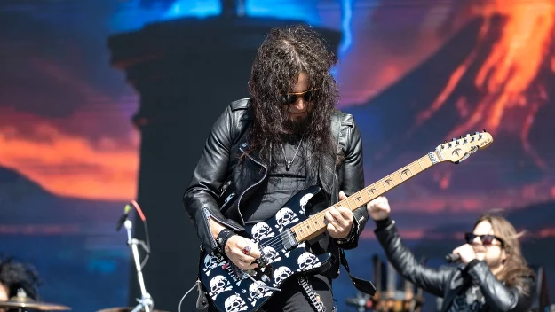 Gitarzysta w skórzanej kurtce grający na elektrycznej gitarze ozdobionej motywem czaszek, towarzyszy mu wokalista z mikrofonem, w tle dynamiczne światła sceniczne.