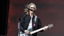 Mężczyzna z długimi włosami gra na gitarze elektrycznej na scenie, śpiewając do mikrofonu; ubrany w czarną kurtkę i okulary przeciwsłoneczne, gitara ozdobiona grafiką.