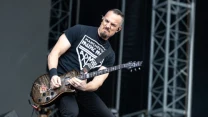 Mężczyzna grający na elektrycznej gitarze na scenie koncertowej, skupiony i dynamiczny wyraz twarzy, ubrany w czarny T-shirt i spodnie.