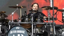 Perkusista grający na rozbudowanym zestawie perkusyjnym ozdobionym logo zespołu Alter Bridge podczas występu scenicznego.