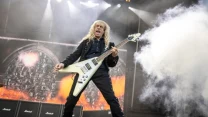 Gitarzysta z długimi blond włosami energicznie grający na scenie podczas koncertu rockowego, w tle ściana wzmacniaczy Marshall i intensywne efekty świetlne oraz dym.