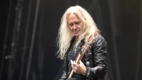 Starszy mężczyzna o długich, siwych włosach grający na gitarze elektrycznej, ubrany w czarną, skórzaną kurtkę i ciemne spodnie, skoncentrowany podczas występu na scenie.