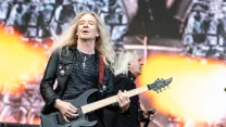 Mężczyzna z długimi blond włosami gra na czarnej gitarze elektrycznej podczas występu na scenie, w tle widoczny kolejny członek zespołu oraz efektowna grafika z motywem ognia.