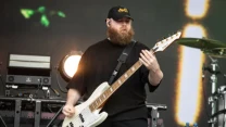 Brodaty mężczyzna w czarnej czapce gra na białej gitarze basowej na scenie koncertowej, w tle sprzęt muzyczny i oświetlenie sceniczne.