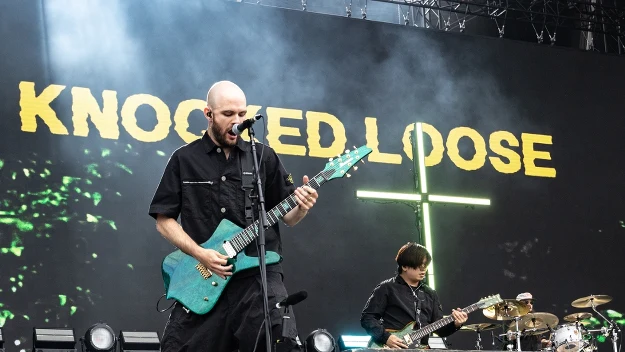 Wokalista grający na gitarze elektrycznej przed dużym neonowym napisem „Knocked Loose”, z basistą i perkusistą na tle profesjonalnej sceny koncertowej.