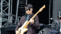 Mężczyzna w ciemnych okularach grający na jasnej gitarze basowej na scenie koncertowej.