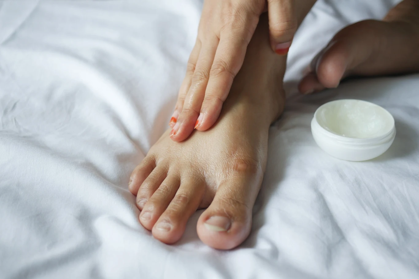 Bare pedicure to postawienie na zadbane stopy w naturalnym wydaniu Dłoń nakłada krem na stopę leżącą na białej pościeli, obok otwarty słoiczek z kosmetykiem.