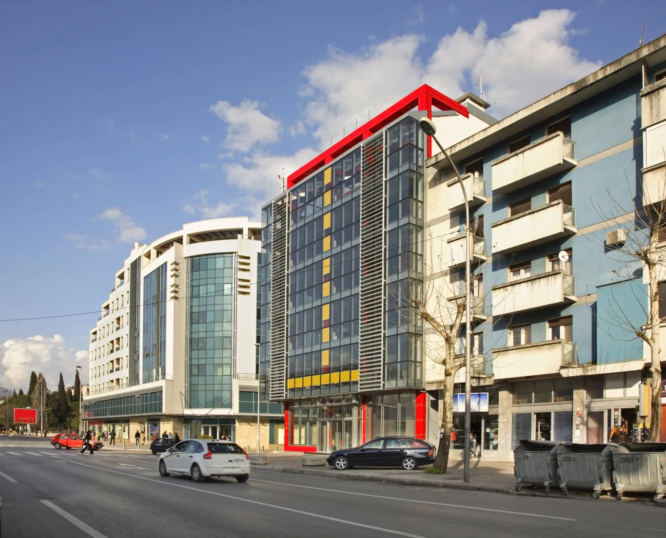 Nowoczesny budynek z przeszkloną fasadą i czerwonymi elementami konstrukcyjnymi w otoczeniu wielopiętrowych bloków mieszkalnych przy ruchliwej ulicy, przy której zaparkowane są samochody.