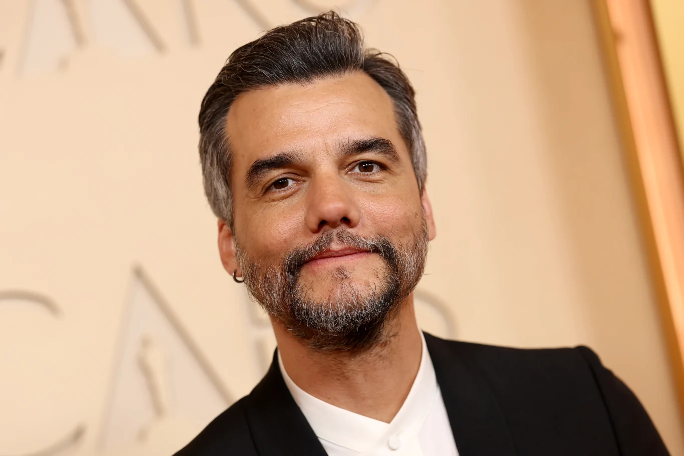 Wagner Moura Wagner Moura w siwych włosach i brodzie, w czarnym garniturze na tle jasnej ścianki z napisami.
