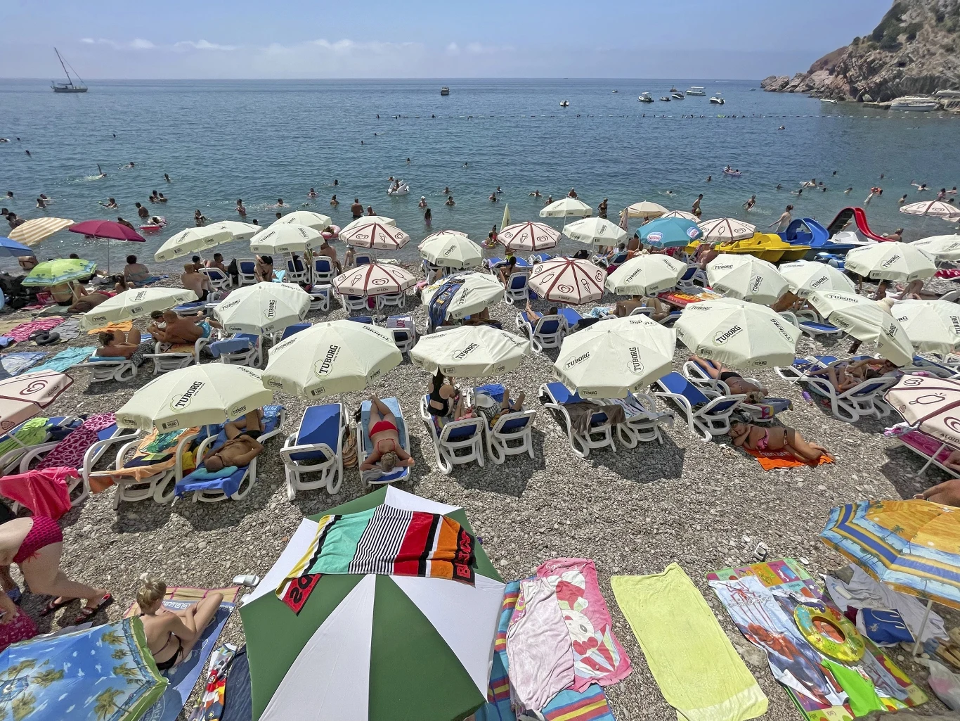 Plaża wypełniona leżakami i parasolami, wokół których wypoczywają ludzie, a w tle spokojne morze z kąpiącymi się osobami i łodziami na wodzie.