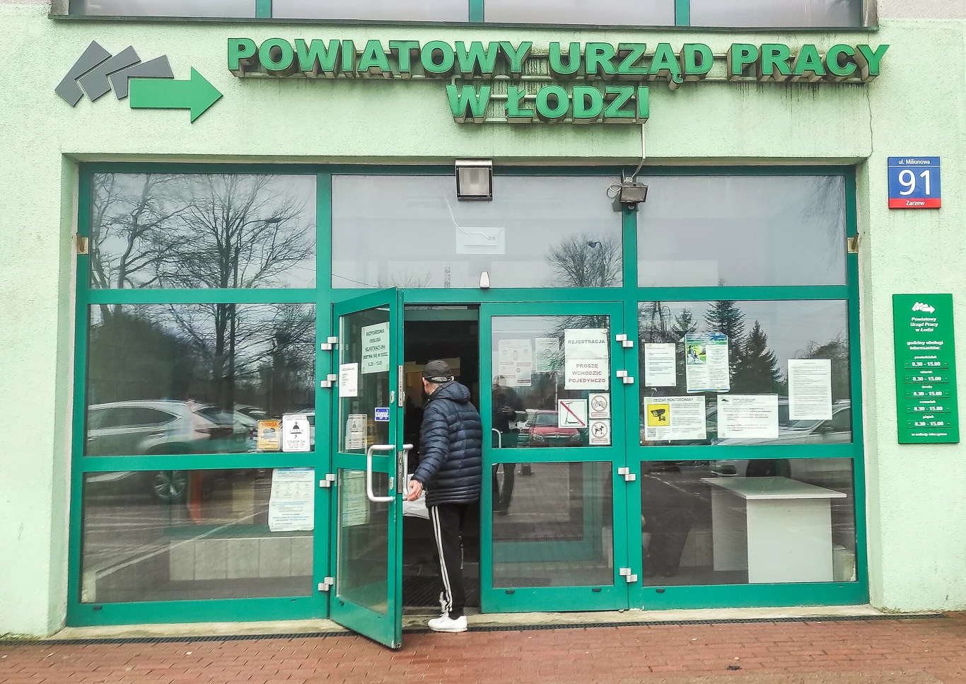 Zasiłek to nie koniec wsparcia dla bezrobotnych. Osoba w ciemnej kurtce i sportowych spodniach wchodzi do Powiatowego Urzędu Pracy w Łodzi; na drzwiach ogłoszenia.