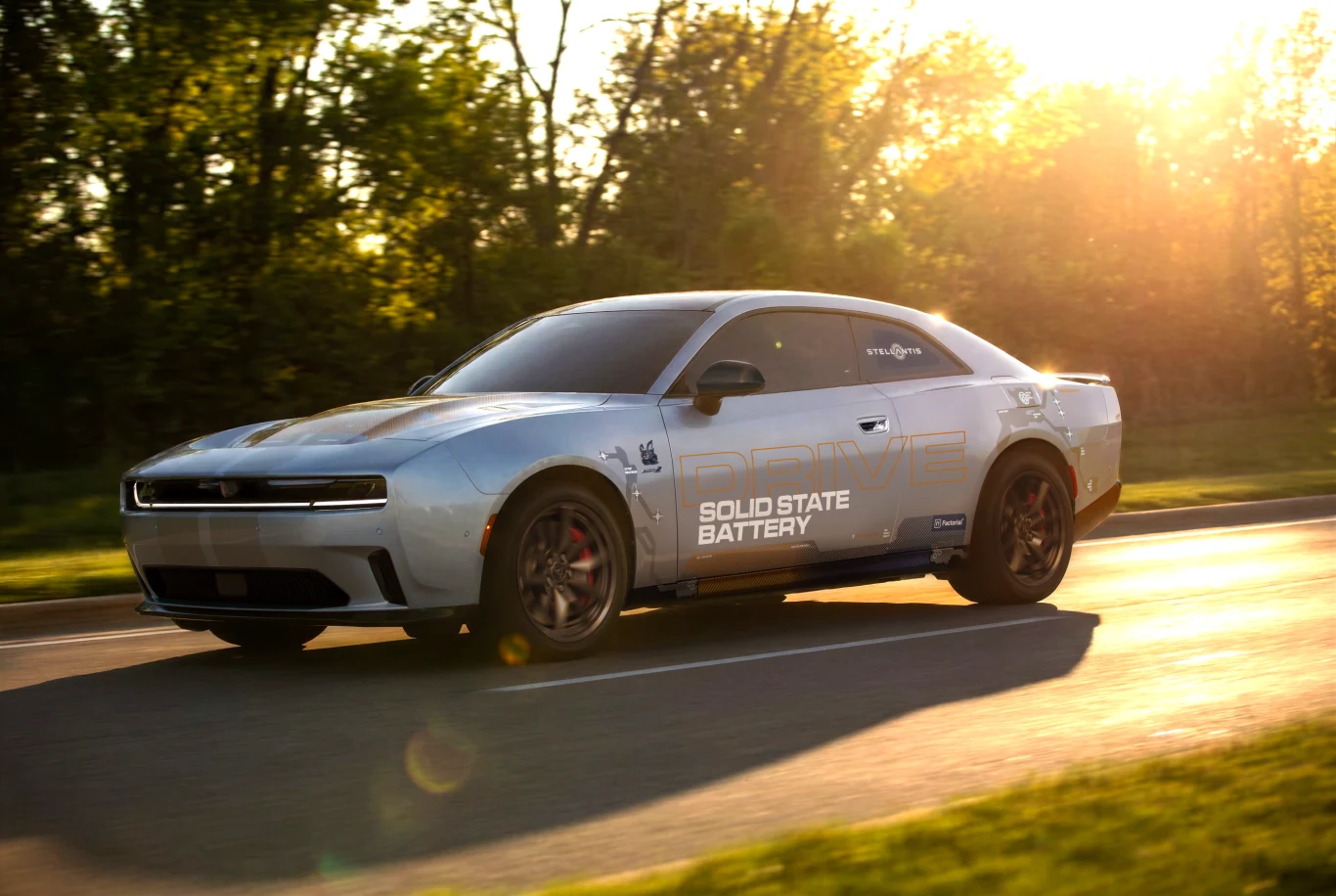 Jasnoszary sportowy samochód z widocznym napisem „Solid State Battery” na drzwiach jedzie asfaltową drogą, otoczony przez drzewa i promienie zachodzącego słońca.