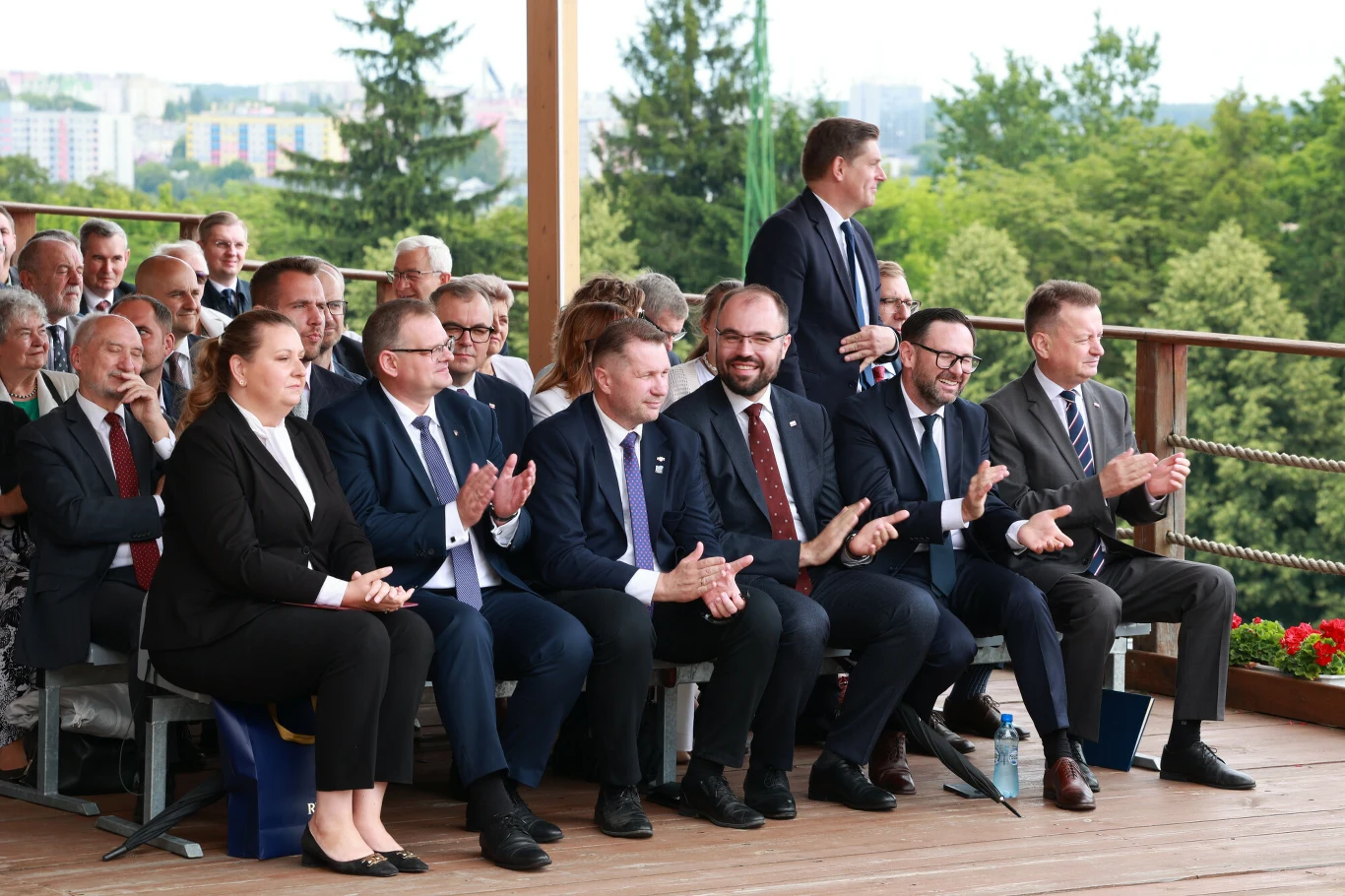 Grupa elegancko ubranych osób siedzących na zadaszonej trybunie, część z nich klaszcze, w tle widoczne zielone drzewa i fragment miasta.