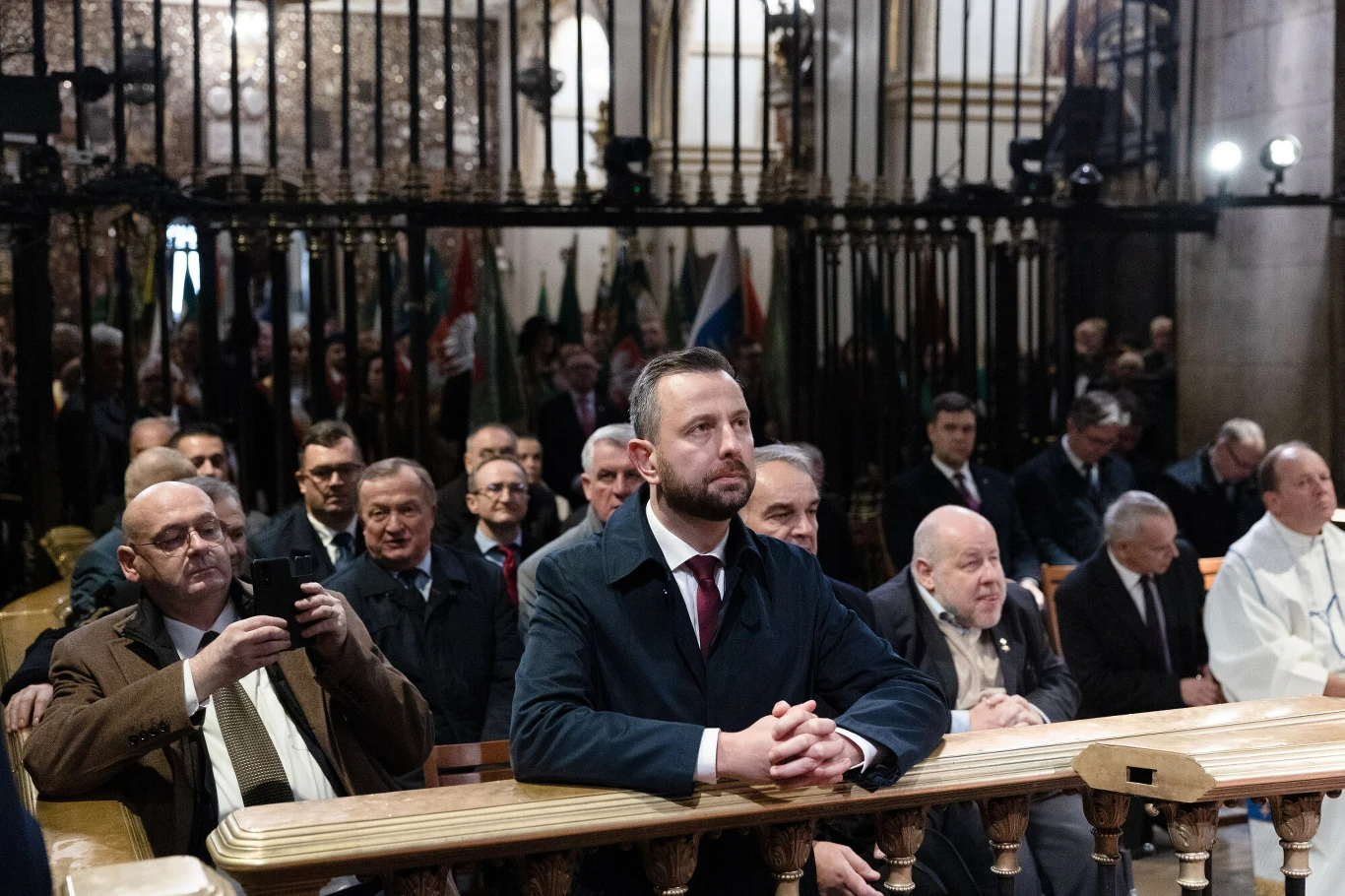 Władysław Kosiniak-Kamysz podczas modlitwy na Jasnej Górze Mężczyzna w garniturze siedzi w pierwszej ławce kościoła, otoczony innymi uczestnikami uroczystości religijnej, w tle widoczne są metalowe kraty oraz grupa osób siedzących i stojących.