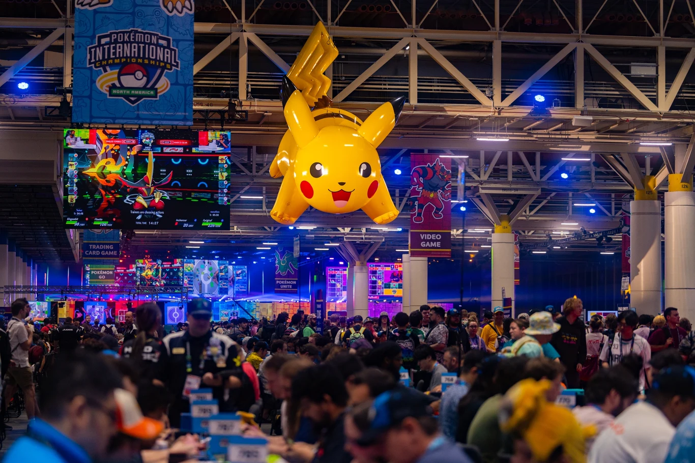 Ogromny, nadmuchiwany Pikachu unoszący się nad tłumem uczestników turnieju Pokémon, kolorowe dekoracje i ekrany z motywami gry w tle.