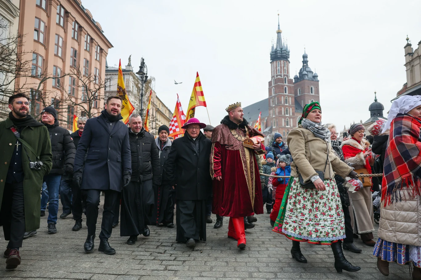 Aleksander Miszalski i kardynał Grzegorz Ryś podczas orszaku Trzech Króli Kolorowy pochód ludzi ubranych w tradycyjne stroje oraz królewskie kostiumy, z flagami w tle, na rynku starego miasta z widoczną bazyliką Mariacką.