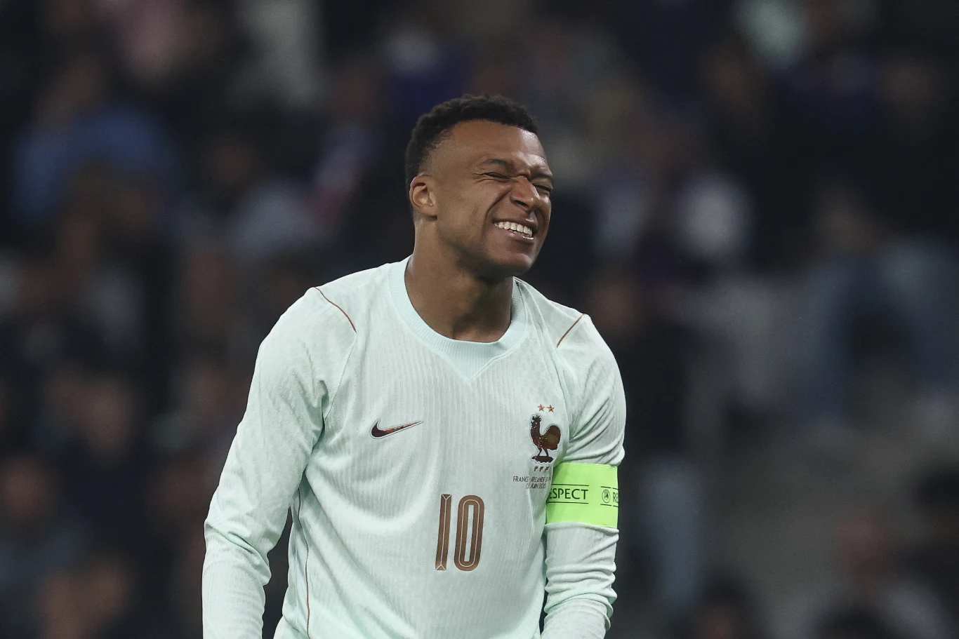 Kylian Mbappe Piłkarz w białym stroju z opaską kapitana na ramieniu, grymaszący z bólu lub rozczarowania, na tle rozmytej publiczności.