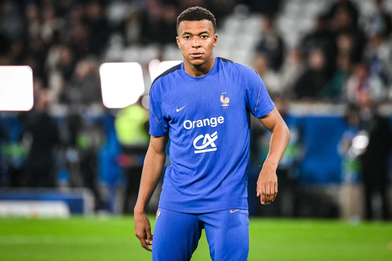 Kylian Mbappe Piłkarz w niebieskim stroju francuskiej reprezentacji stoi na murawie stadionu, skupiony, z lekko zgiętą ręką i delikatnym wyrazem twarzy.