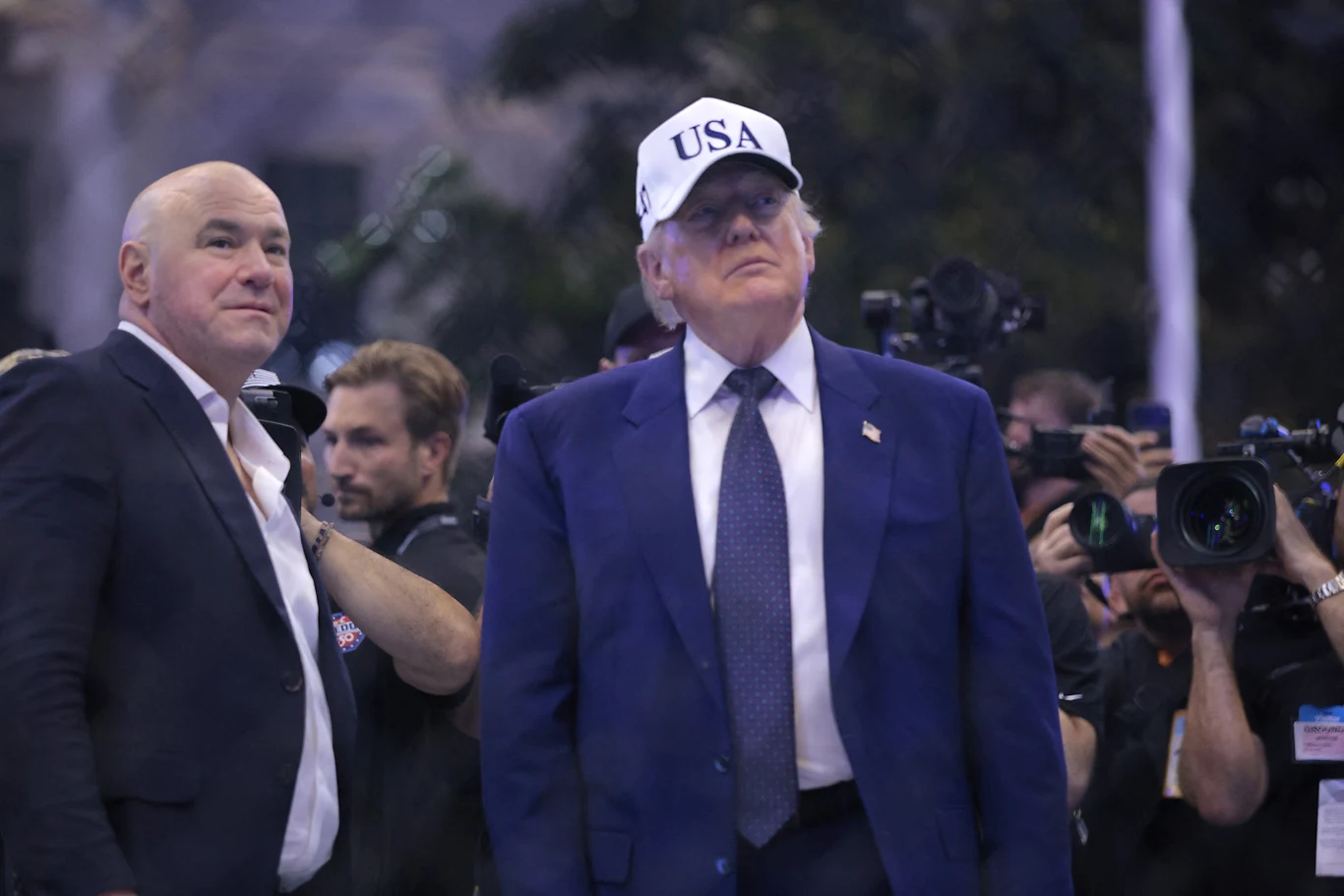 Dwóch mężczyzn w garniturach na pierwszym planie, jeden z nich w białej czapce z napisem USA, otoczeni przez grupę fotografów z aparatami.