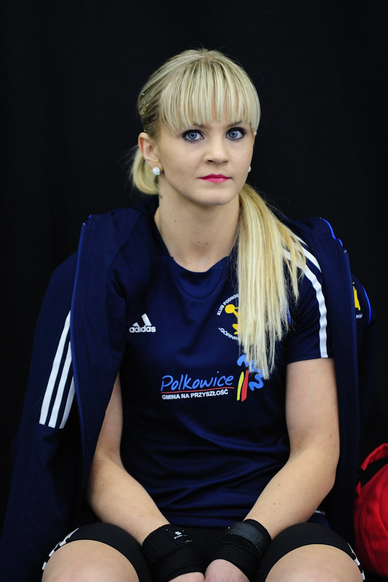 Kobieta w sportowej koszulce z logo klubu i napisem Polkowice, siedząca z rękami opartymi na kolanach, z blond włosami spiętymi w koński ogon, na tle ciemnego tła.
