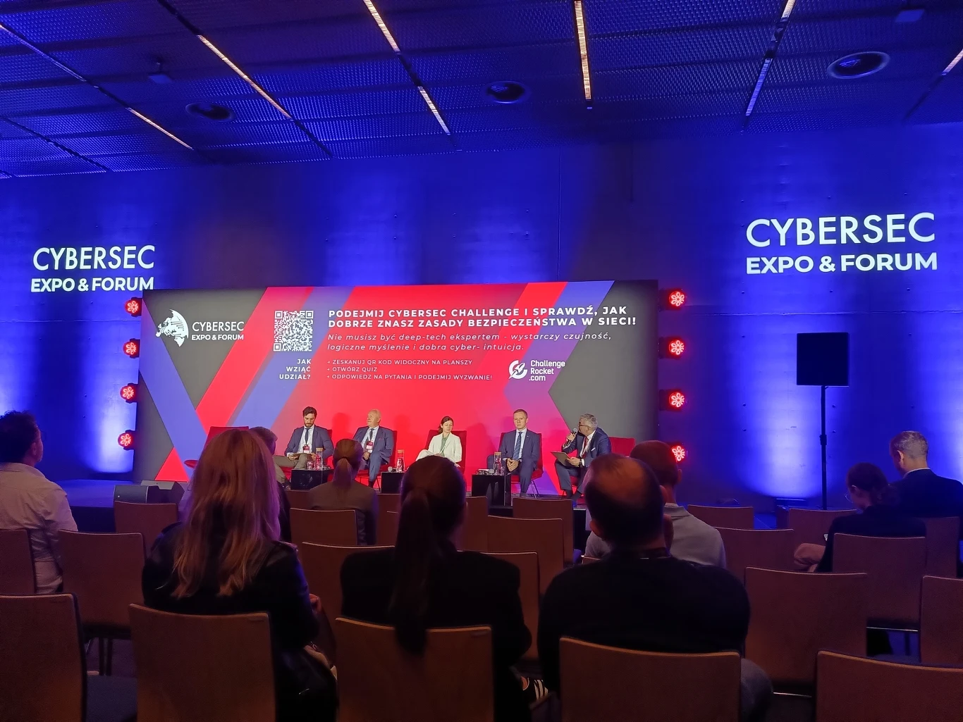 Eksperci debatowali nad możliwościami aktywnej obrony Polski i NATO przed agresją Rosji w cyberprzestrzeni Panel ekspertów siedzących na scenie podczas konferencji CYBERSEC Expo & Forum, z publicznością obserwującą dyskusję i nowoczesnym tłem podświetlonym na fioletowo-niebiesko.