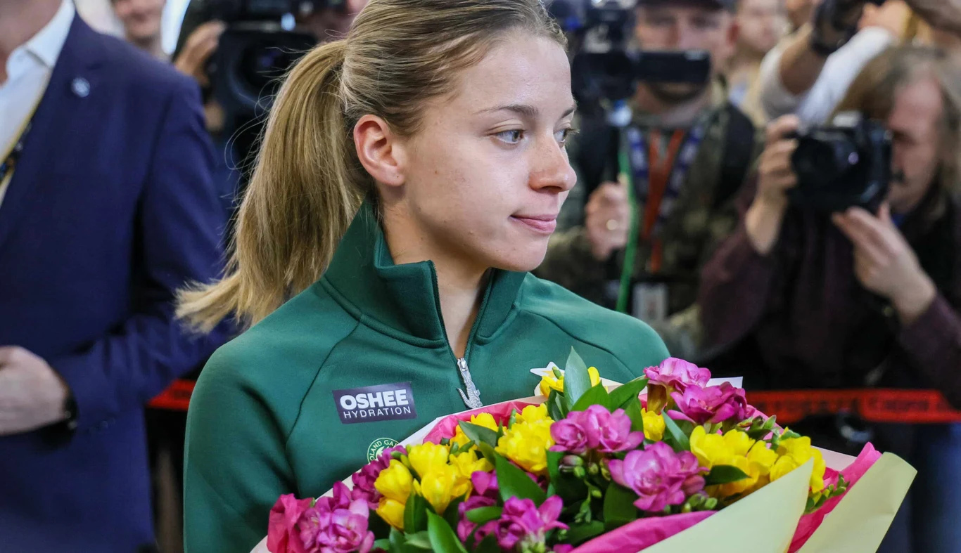Maja Chwalińska Młoda kobieta w zielonej sportowej kurtce trzyma duży bukiet kolorowych kwiatów, wokół niej stoją fotografowie i dziennikarze z aparatami.