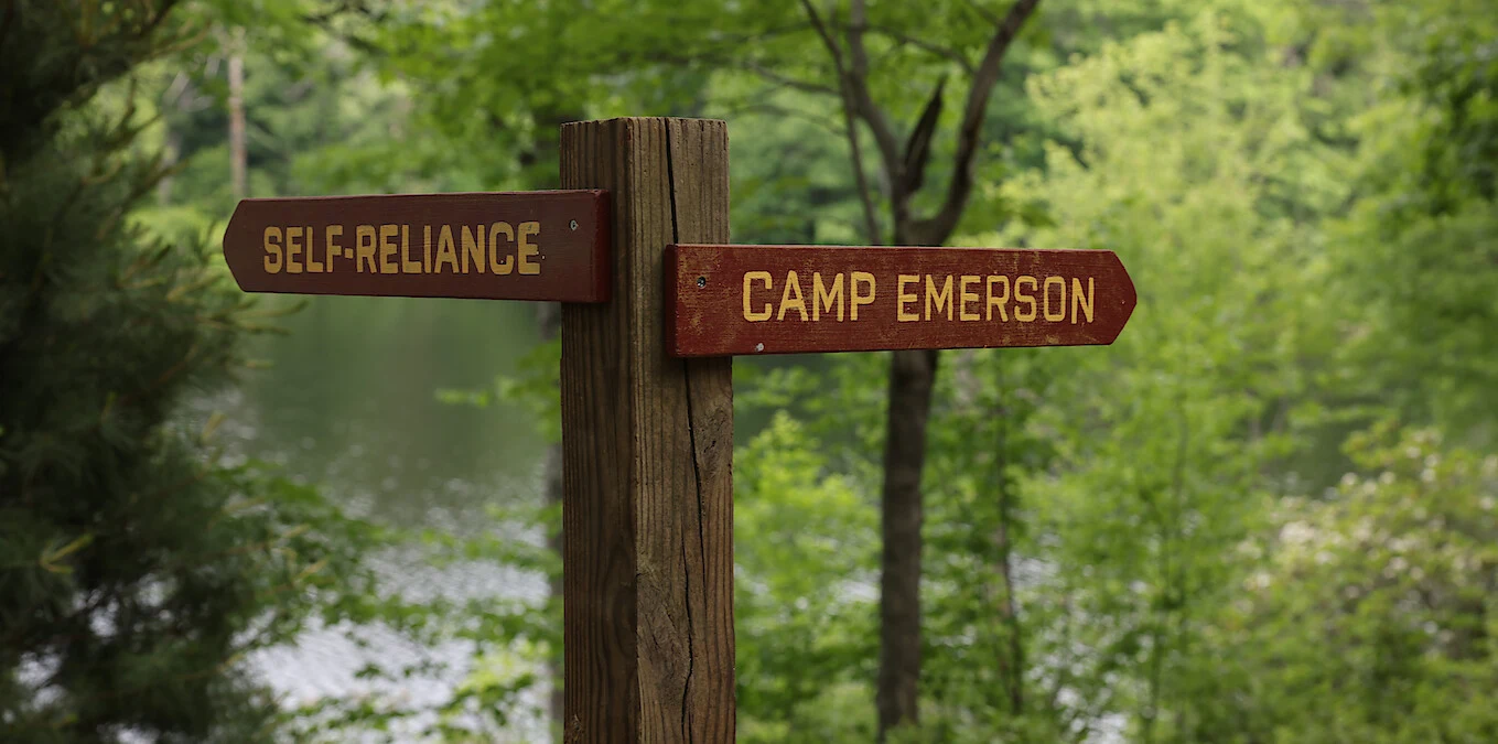 Drewniany słupek z dwoma brązowymi drogowskazami wskazującymi kierunki: jeden z napisem „SELF-RELIANCE”, drugi „CAMP EMERSON”, otoczony zielonymi drzewami i widokiem na jezioro w tle.