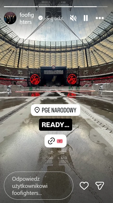 Widok na pustą płytę stadionu PGE Narodowy z centralnie ustawioną dużą sceną koncertową, na której widoczne są dwa czerwone logotypy zespołu Foo Fighters po obu stronach ekranu głównego.