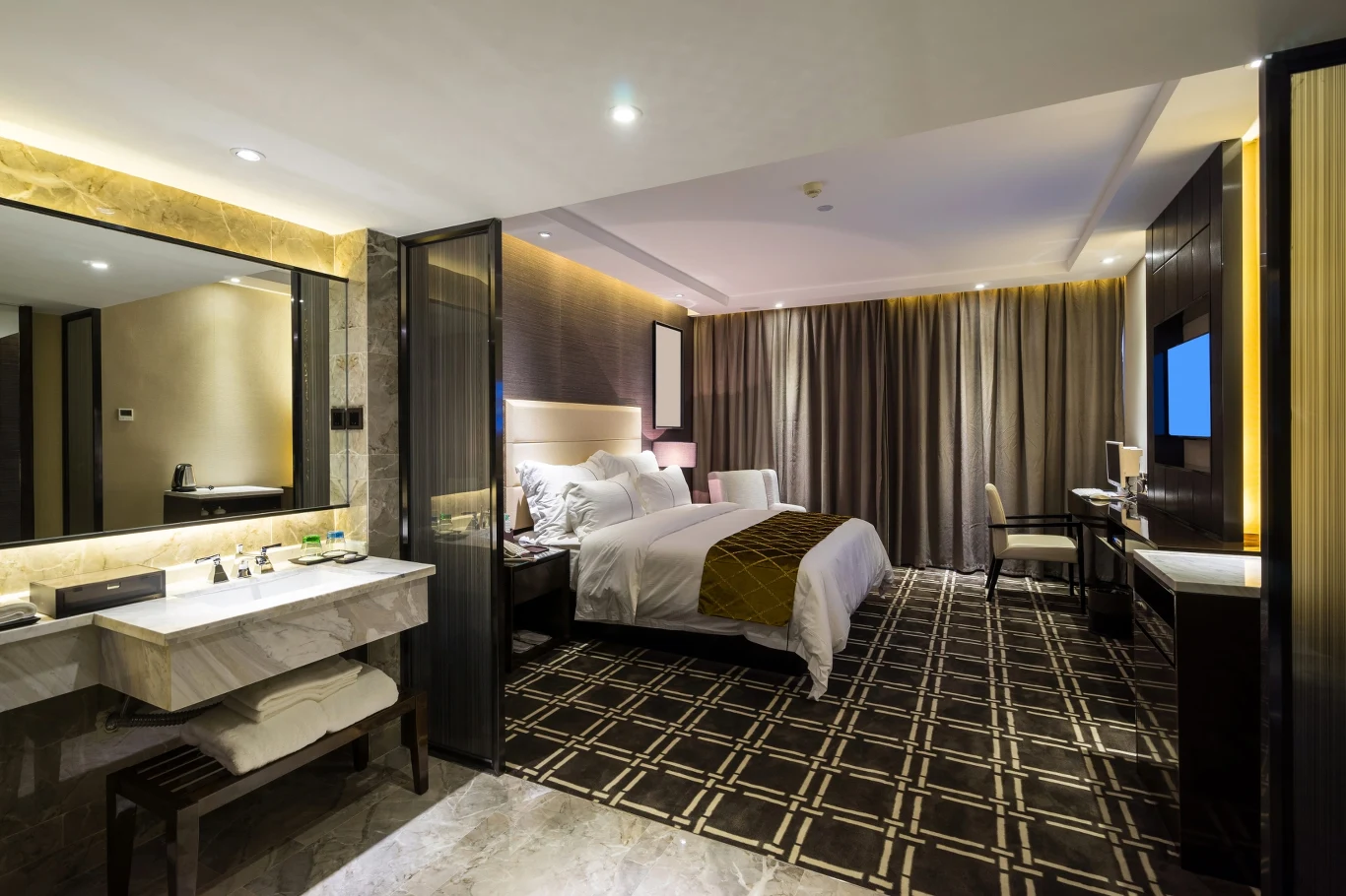 Elegancki pokój hotelowy z dużym łóżkiem, biurkiem z krzesłem, telewizorem na ścianie oraz przestronną łazienką z lustrem i umywalką po lewej stronie.