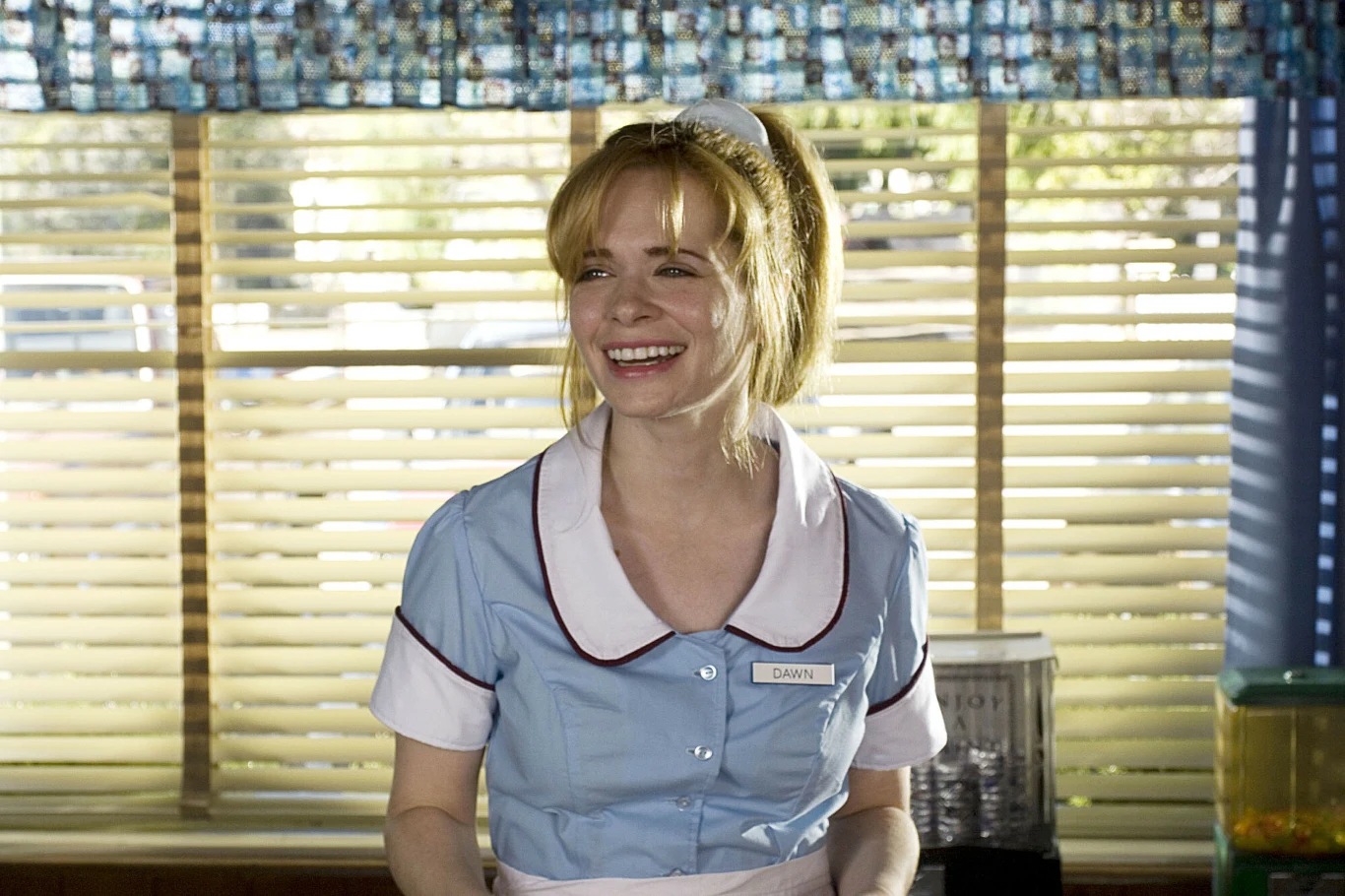 Adrienne Shelly w filmie "Kelnerka" Kobieta w jasnoniebieskim uniformie kelnerki z białym kołnierzykiem i przypiętą plakietką imienną, uśmiechnięta, stojąca przy oknie z żaluzjami.