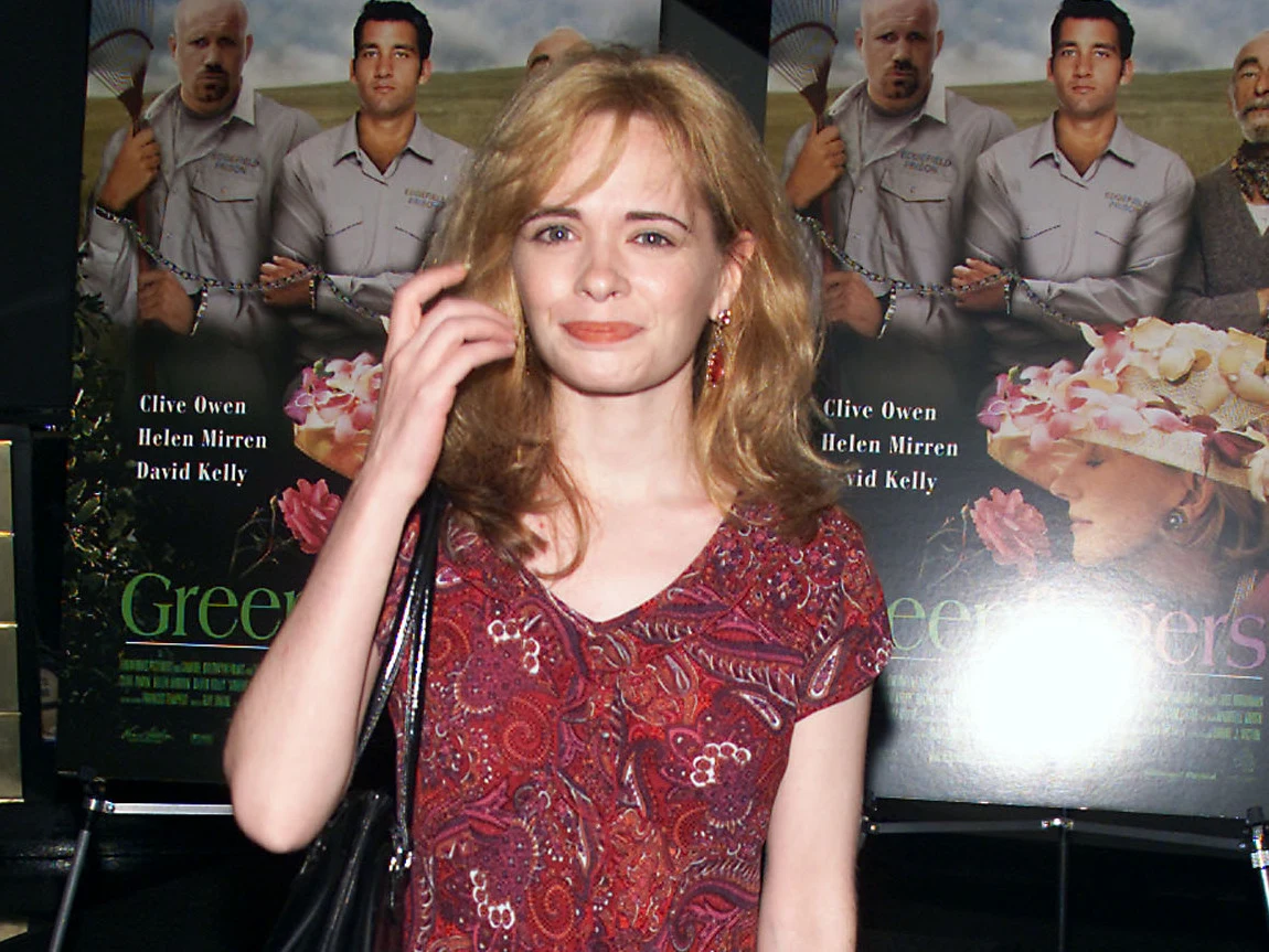 Adrienne Shelly w 2001 roku Kobieta w czerwonej bluzce z jasnymi włosami stoi na tle dwóch plakatów filmowych z widocznymi twarzami aktorów i nazwiskami Clive Owen, Helen Mirren oraz David Kelly.