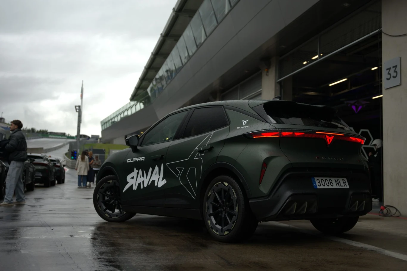 Raval to najnowszy model w gamie Cupry - skierowany do klientów szukających taniego elektryka, a przy tym auta dostarczającego przyjemnych wrażeń z jazdy. Nowoczesny SUV Cupra z charakterystycznymi czerwonymi tylnymi światłami i dużym napisem SAVAL na bokach, stojący na mokrej nawierzchni przy wejściu do boksu toru wyścigowego.