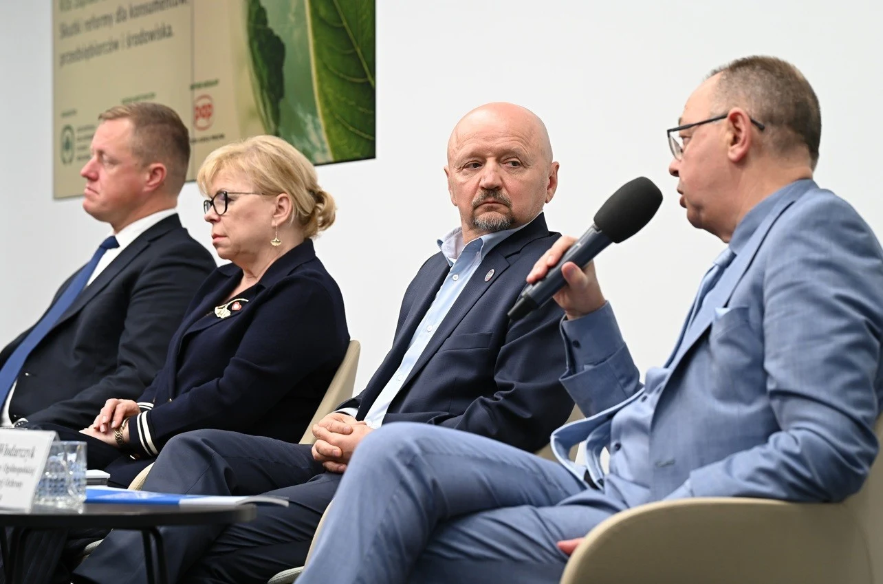 Czworo elegancko ubranych osób siedzi obok siebie na panelu dyskusyjnym, jedna z nich trzyma mikrofon i mówi, pozostali słuchają z uwagą.