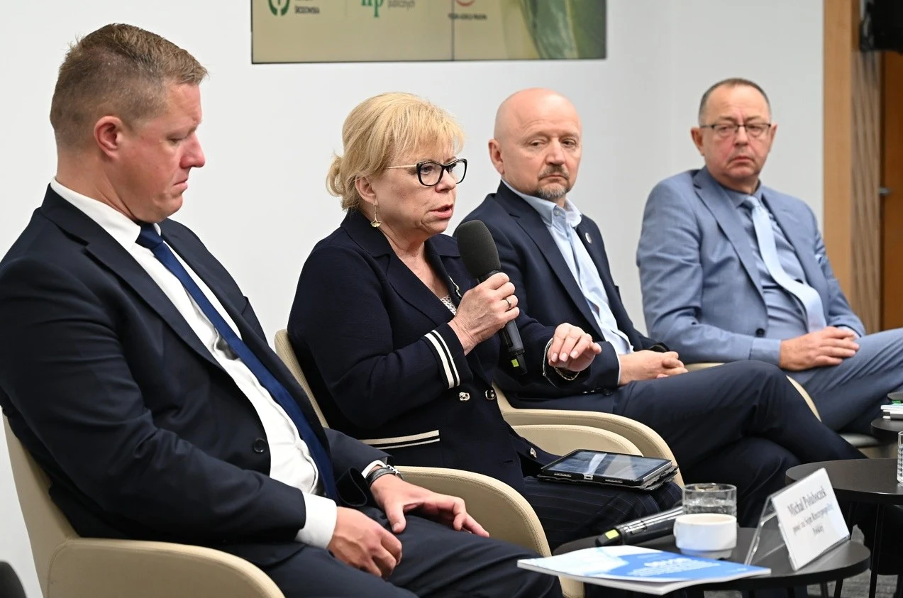 Cztery osoby w garniturach siedzą w rzędzie podczas panelu dyskusyjnego, jedna z kobiet trzyma mikrofon i mówi, pozostali słuchają, na stoliku przed nimi widoczne są notatki i szklanka z wodą.