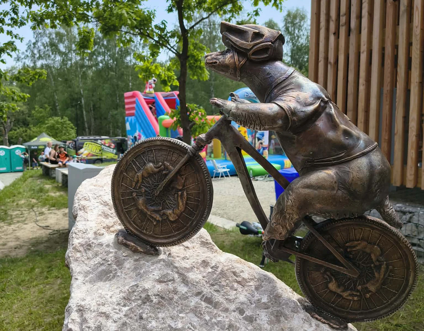 Miś Rowerzysta w Polanicy - Zdroju Brązowa rzeźba szczura w stroju rowerzysty jadącego na rowerze, ustawiona na kamieniu, w tle kolorowe dmuchańce oraz drzewa.