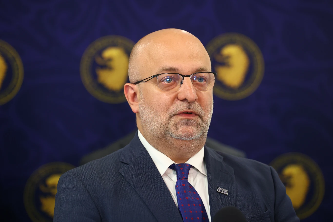 Łysy mężczyzna w okularach, ubrany w ciemny garnitur i krawat, stoi przed ciemnoniebieskim tłem z żółtymi emblematami lwów.