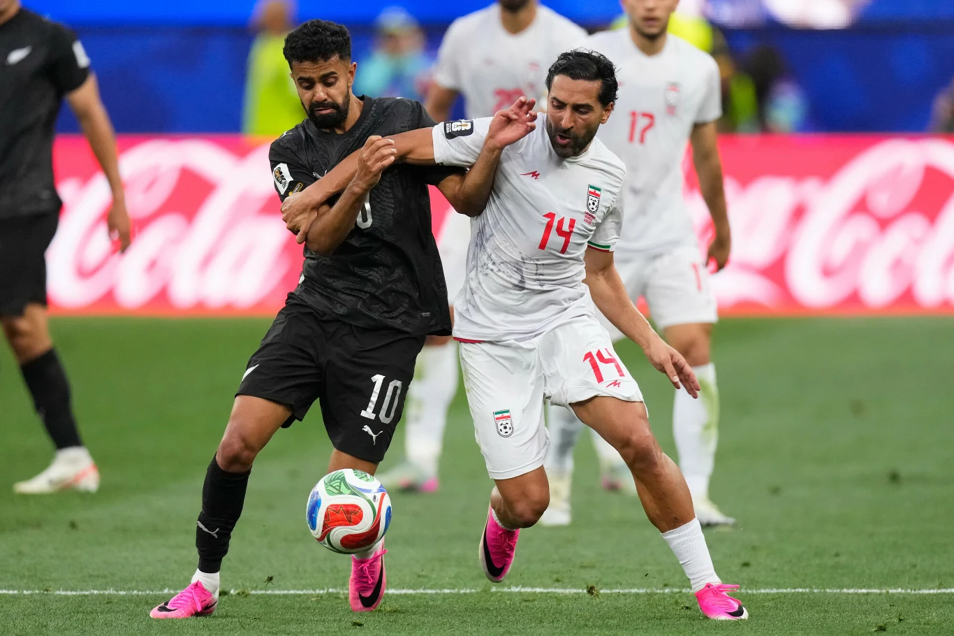 Sarpreet Singh i Saman Ghoddo, Iran - Nowa Zelandia na MŚ 2026 Dwóch piłkarzy rywalizuje o piłkę podczas meczu, jeden z nich ubrany jest w czarny strój z numerem 10, drugi w biały z numerem 14.