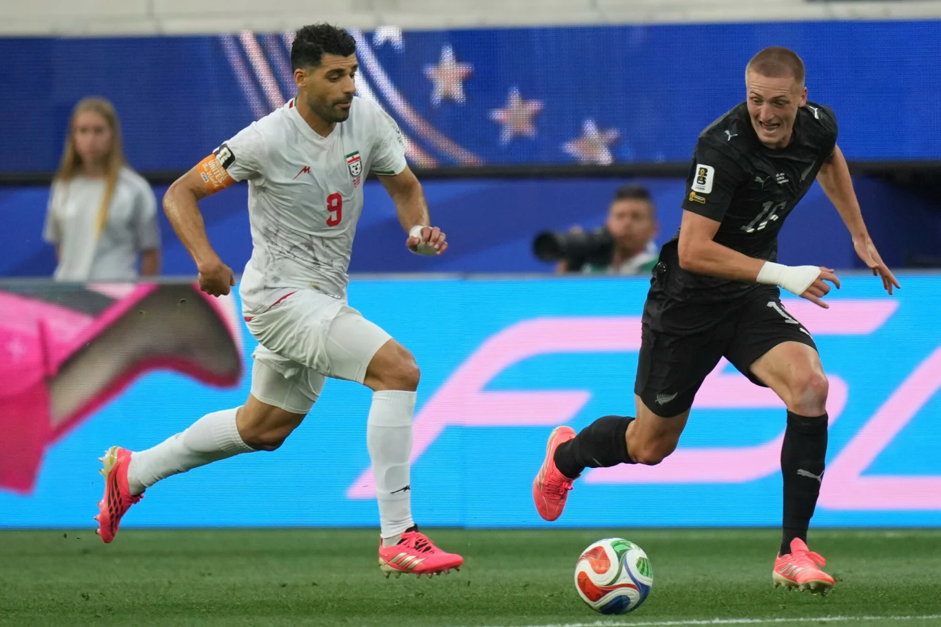 Mehdi Taremi i Finn Surman, Iran - Nowa Zelandia na MŚ 2026 Dwóch piłkarzy rywalizuje o piłkę podczas meczu; zawodnik w jasnym stroju biegnie obok przeciwnika w czarnym stroju, koncentrując się na walce o kontrolę nad futbolówką.